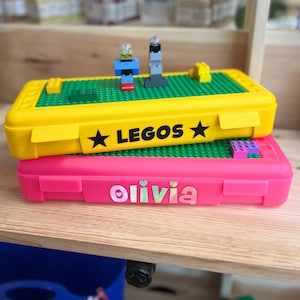 May include: Two yellow and pink plastic storage cases with a Lego base. The yellow case has a black star on each side and the text "LEGOS" in black. The pink case has a sticker with the name "olivia" in rainbow letters.