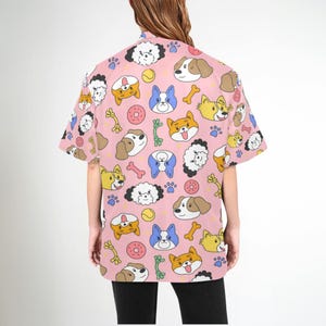 Pu&ograve; includere: Una camicia rosa a maniche corte con bottoni e un motivo ripetitivo di cani dei cartoni animati, ossa e giocattoli per cani.