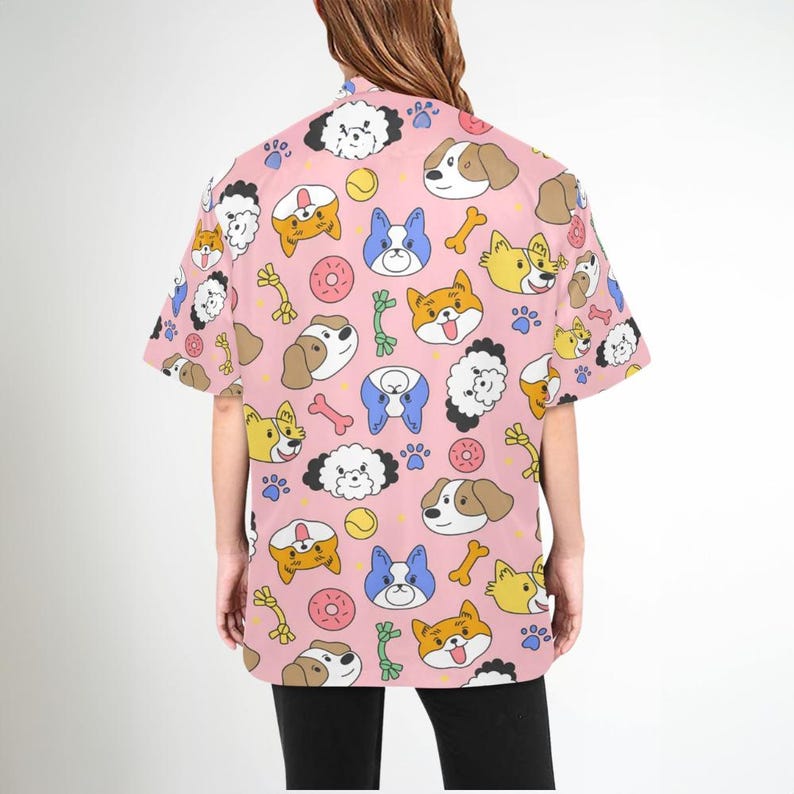Pu&ograve; includere: Una camicia rosa a maniche corte con bottoni e un motivo ripetitivo di cani dei cartoni animati, ossa e giocattoli per cani.