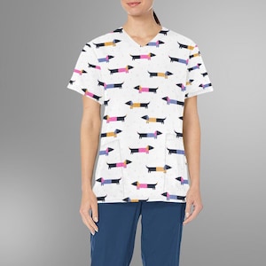 May include: White V-neck scrub top with a repeating pattern of colorful dachshund dogs. The dogs are in various colors, including pink, blue, yellow, and black. The top has two front pockets and short sleeves. The model is wearing blue pants.