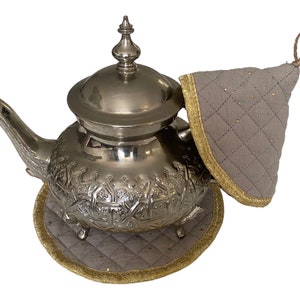 May include: Silver teapot with intricate designs and a grey quilted tea cosy. The teapot has a round body and a spout on the side. The tea cosy has a gold trim.