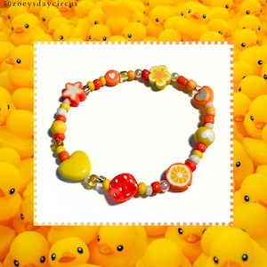 Könnte beinhalten: Ein farbenfrohes Perlenarmband mit verschiedenen Charms, darunter ein Herz, ein Stern, eine Blume, ein Würfel und eine Orangenscheibe. Die Perlen sind gelb, orange, rot und weiß.