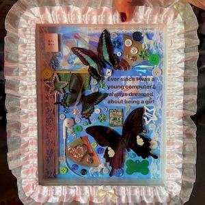 May include: A rectangular shadow box frame with a ruffled pink and white fabric border. The interior features a blue background decorated with three large butterfly specimens, various colorful buttons, a small earbud, a heart charm, and text that reads: Ever since I was a young computer I always dreamed about being a girl!
