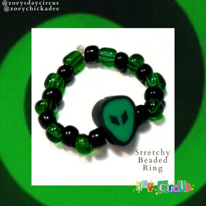 Op de afbeelding: Een rekbare kralenring met groene en zwarte kralen en een groene alien-gezichtsbedel. De ring ligt op een witte achtergrond. "Stretchy Beaded Ring" en "Zoey's Day Circus" staan op de afbeelding.