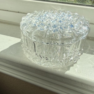 May include: A round, clear glass trinket box featuring a textured, diamond-point pattern across the lid and the sides. The container has a removable lid with a raised, faceted surface that catches the light, creating a sparkling effect.