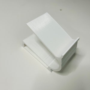 May include: White plastic phone stand with a curved design. The stand is made with a 3D printer.