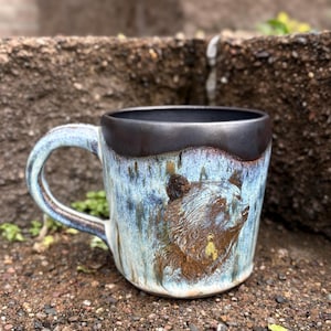May include: A handmade ceramic mug with a bear design. The mug features a blue and brown glaze with a dark brown rim and handle. The bear is sculpted in relief on the side of the mug.