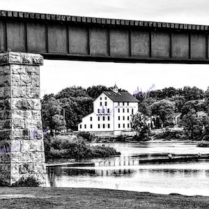 Puede incluir: Fotografía en blanco y negro de un pilar de puente de piedra y una estructura de puente metálico. Un edificio blanco con muchas ventanas es visible en el fondo, junto con árboles y un río. El edificio tiene texto.