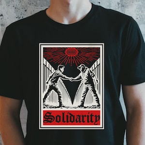 May include: Black t-shirt featuring a graphic print of two figures shaking hands in front of a fence. The graphic is in black and white with red accents. The text "Solidarity" is printed below the graphic.
