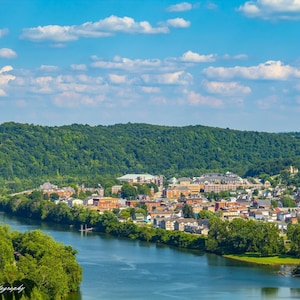 Könnte beinhalten: Eine Luftaufnahme einer Stadt, die zwischen einem Fluss und einem grünen Hügel eingebettet ist. Die Stadt ist voller Gebäude, Bäume und einem gewundenen Fluss. Der Himmel ist blau mit weißen Wolken.