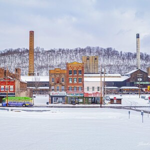 May include: A winter scene of a small town with snow-covered streets and buildings. A colorful bus is parked in front of the buildings. A tall brick smokestack and a snow-covered hill are in the background.