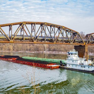 May include: A white tugboat pulling a barge loaded with black coal under a rusty metal bridge. The barge is green and red with a black cargo.