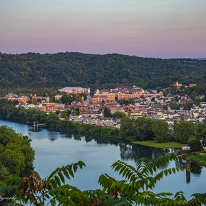 Puede incluir: Una vista aérea de una ciudad con un río serpenteando a través de ella. La ciudad está ubicada en un valle con exuberantes árboles verdes y un cielo rosado en el fondo.