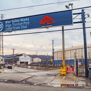 Może przedstawiać: Wejście do Mon Valley Works, Edgar Thomson Plant, United States Steel. Duży znak głosi "Mon Valley Works" i "United States Steel". Pomnik mężczyzny w pomarańczowych spodniach i niebieskiej koszuli stoi przy wejściu.