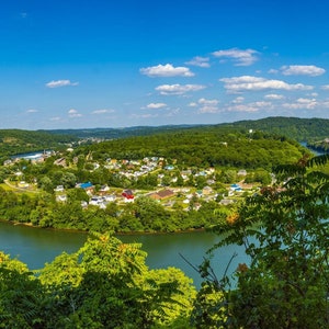 Puede incluir: Vista aérea de un pequeño pueblo ubicado en una curva de un río, rodeado de exuberantes árboles verdes y un cielo azul con nubes blancas.