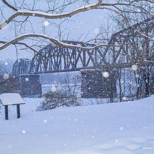 May include: A winter landscape featuring a snow-covered bridge with a metal truss design. The scene includes a sign, trees, and falling snowflakes, creating a serene, cold atmosphere. The image is a photograph.