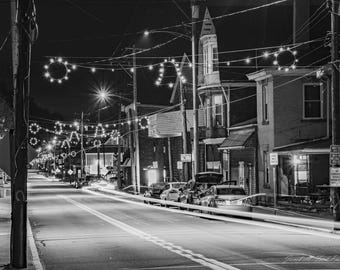 May include: A black and white photo of a street lined with buildings at night. The street is decorated with string lights. There are cars parked on the side of the street.
