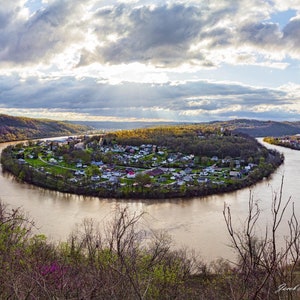 Puede incluir: Una vista panorámica de una ciudad ubicada en una curva de un río. El río es ancho y marrón, y la ciudad está rodeada de árboles verdes exuberantes. El cielo está nublado con rayos de sol que atraviesan las nubes.