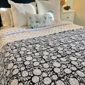 May include: A bed featuring a navy blue, button-tufted headboard and white bedding. The bed is dressed with white pillows and a decorative pillow with a blue floral design. A black and white floral quilt covers the bed's foot.