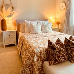 May include: A bedroom with a white bed frame and a brown floral patterned quilt. The bed has white pillows and a brown patterned throw pillow. There is a white nightstand with two drawers on the left side of the bed and a white nightstand with one drawer on the right side of the bed. There is a white lamp on each nightstand. There is a white rug on the floor. There is a white wall behind the bed. There is a white ceiling above the bed. There is a white window on the right side of the bed. There is a white curtain on the window. There is a white door on the left side of the bed. There is a white door frame around the door. There is a white trim around the window. There is a white trim around the door. There is a white trim around the ceiling. There is a white trim around the floor. There is a white trim around the wall. There is a white trim around the bed. There is a white trim around the nightstand. There is a white trim around the lamp. There is a white trim around the rug. There is a white trim around the curtain. There is a white trim around the door frame. There is a white trim around the window frame. There is a white trim around the ceiling frame. There is a white trim around the floor frame. There is a white trim around the wall frame. There is a white trim around the bed frame. There is a white trim around the nightstand frame. There is a white trim around the lamp frame. There is a white trim around the rug frame. There is a white trim around the curtain frame. There is a white trim around the door frame frame. There is a white trim around the window frame frame. There is a white trim around the ceiling frame frame. There is a white trim around the floor frame frame. There is a white trim around the wall frame frame.