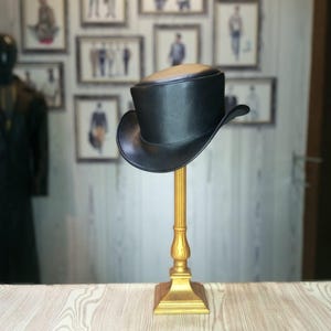 May include: A black top hat with a curved brim is displayed on a gold-colored stand. The hat has a smooth texture and a rounded crown. The stand has a square base and a decorative, ornate design. The background features framed pictures.