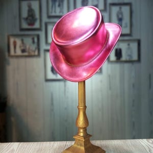 May include: A pink top hat with a shiny finish sits on a gold stand. The hat is on a wooden surface with a blurred background of framed pictures.