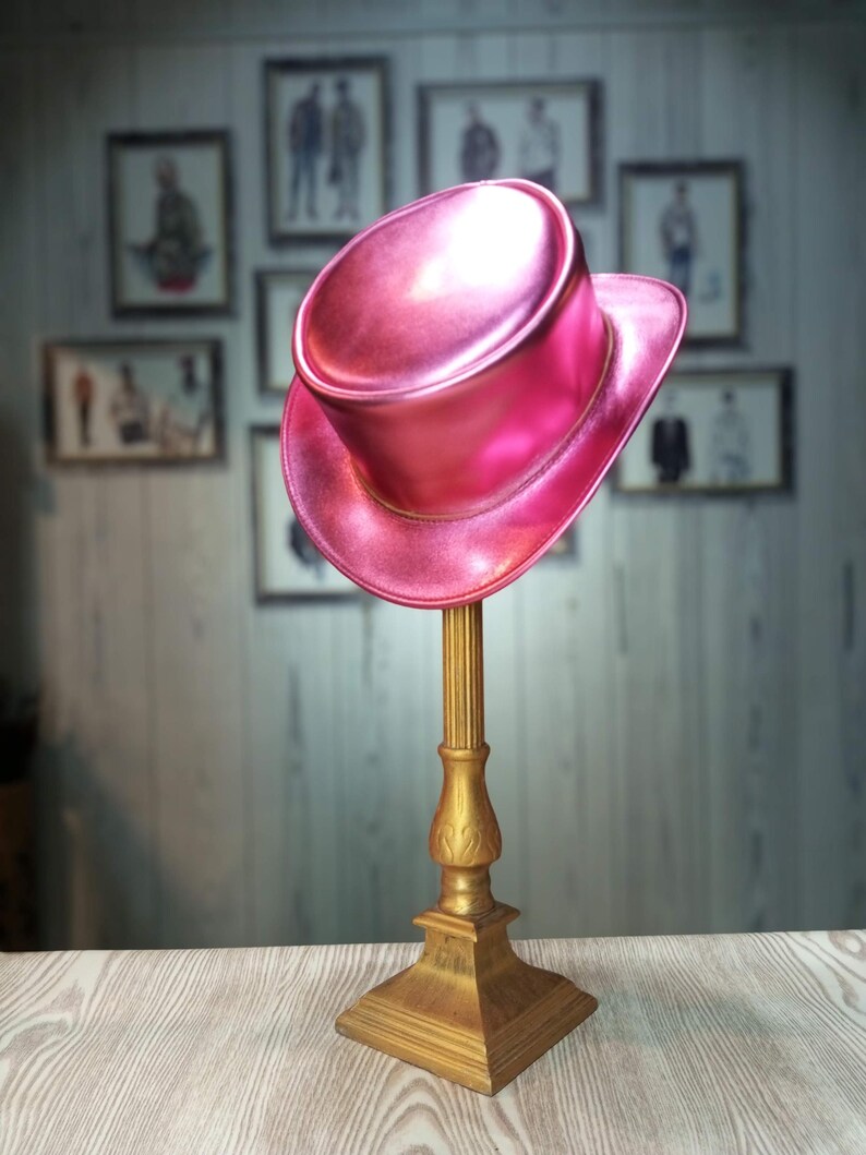May include: A pink top hat with a shiny finish sits on a gold stand. The hat is on a wooden surface with a blurred background of framed pictures.