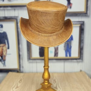 May include: A brown leather top hat displayed on a gold-colored stand. The hat has a rounded crown and a wide brim. The stand is ornate with a square base and a decorative column.