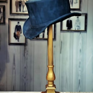 May include: A dark blue top hat rests on a gold-colored hat stand. The stand has a decorative base and a tall, slender stem. The background features framed pictures of people in suits, creating a vintage aesthetic.
