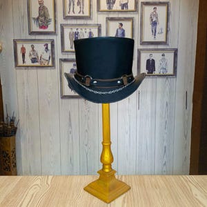 May include: A black leather top hat with a chain detail and a gold stand. The hat is on a wooden surface.