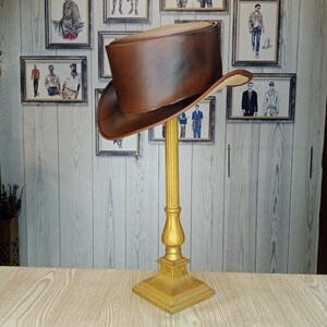 May include: A brown leather top hat displayed on a gold-colored stand. The hat has a wide brim and a tall, cylindrical crown. The stand is ornate, with a square base and a fluted column. The background features framed illustrations.