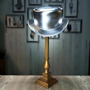 May include: A silver top hat with a wide brim sits on a gold metal stand. The hat is on a wooden surface.