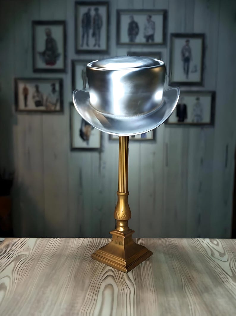May include: A silver top hat with a wide brim sits on a gold metal stand. The hat is on a wooden surface.