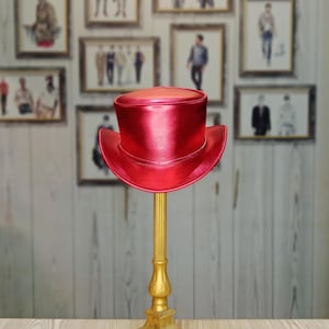 May include: A bright red top hat with a shiny finish sits on a gold stand. The hat is made of leather and has a wide brim.