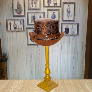 Può includere: Un cappello a cilindro marrone e nero in pelle di serpente con un bordo marrone e dettagli dorati. Il cappello è esposto su un supporto dorato.