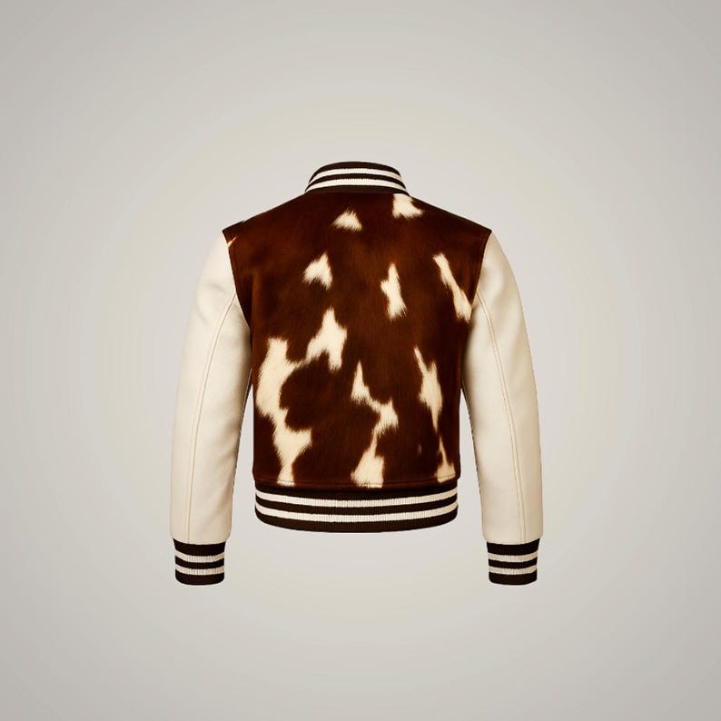 Op de afbeelding: Een varsity-jack met een bruin en wit koeienhuidpatroon. De jas heeft cr&egrave;mekleurige mouwen en een geribbelde kraag, manchetten en zoom met zwart-witte strepen. De achterkant van de jas is zichtbaar.