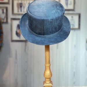 May include: A blue top hat with a wide brim sits on a gold-colored decorative stand. The hat has a slightly distressed appearance. The stand is ornate, with a square base and a fluted column.