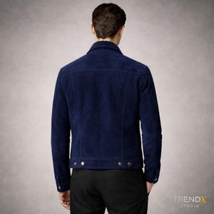 May include: A navy blue suede jacket with a classic collar and buttoned cuffs. The jacket has two chest pockets and a button closure. The model is wearing black trousers. A stylish outerwear piece.