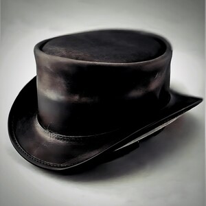 May include: A black leather top hat with a rounded crown and a wide, slightly curved brim. The hat has a classic design, with a smooth, polished finish. The hat is a formal accessory.
