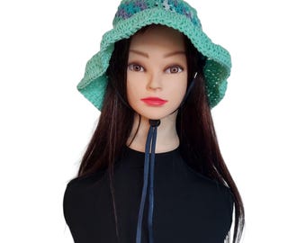 May include: A handmade crochet sun hat in shades of teal, blue, and purple. The hat features a wide brim and a drawstring for an adjustable fit. The hat is displayed on a mannequin head with long, dark hair.