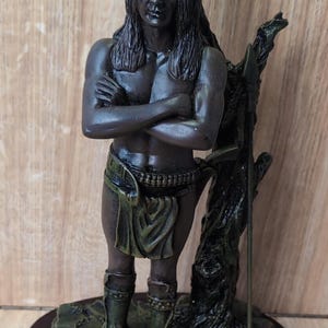 May include: A dark bronze-toned figurine of a person with long hair and a headband, standing with arms crossed. The figure wears a loincloth and leg guards, positioned next to a tree trunk with a spear. A plaque reads The Juliana Collection.