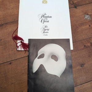 May include: A white programme booklet featuring a gold crest and the text The Phantom of the Opera Her Majesty's Theatre London with a red tassel, alongside a black booklet displaying a white mask graphic and the text Her Majesty's Theatre.