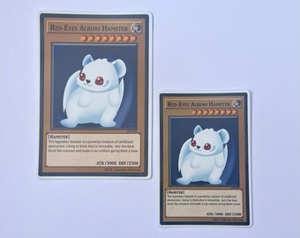 Könnte beinhalten: Zwei Sammelkarten mit einem weißen Hamster mit roten Augen. Die Karten sind braun und goldfarben mit dem Text "Red-Eyes Albino Hamster" und "[HAMSTER]" darauf. Der Text auf den Karten beschreibt den Hamster als eine mächtige Kreatur aus Pappe, die Zerstörung anrichtet.