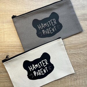May include: Two zippered pouches with a black silhouette of a hamster with the text "Hamster Parent" in white. One pouch is gray and the other is white.