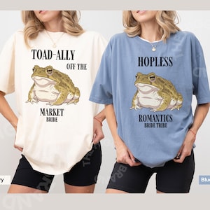May include: Two oversized t-shirts, one ivory and one blue jean, each featuring a cartoon toad illustration. The ivory shirt reads "TOAD-ALLY OFF THE MARKET BRIDE." The blue jean shirt reads "HOPLESS ROMANTICS BRIDE TRIBE."