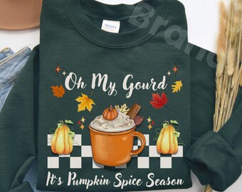 May include: Forest green sweatshirt with the text "Oh My Gourd" and "It's Pumpkin Spice Season". The design includes a pumpkin spice latte, pumpkins, and fall leaves. The sweatshirt is displayed with a brown hat and blue jeans.