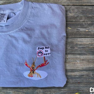 May include: A light gray Comfort Colors t-shirt with an embroidered design. The design features a red crawfish holding a sign that reads "Say NO to POT!" The shirt is folded on a wooden surface.