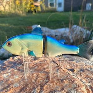 May include: A blue and white fishing lure with a realistic fish design. The lure has a jointed body and a tail with a blue and white color scheme. The lure is on a display stand.