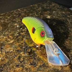 May include: A fishing lure with a green and yellow textured body, a dark purple back, and a red eye. It features a clear plastic diving lip at the front and two sharp metal treble hooks attached to the underside of the body for catching fish.