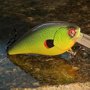 May include: A green and yellow fishing lure with a textured scale pattern, a black spot with a red accent on the side, a red eye, and a clear plastic diving lip. The lure features two sharp metal treble hooks attached to the underside and rear.